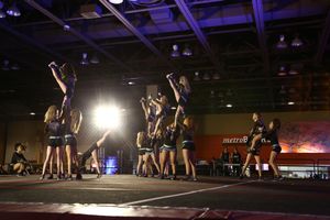 A cheer squad performs a routine with flyers raised overhead as teammates stand in formation on a competition stage.