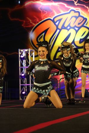 Smiling cheerleader in a black and teal uniform kneels on stage while teammates stand behind her.