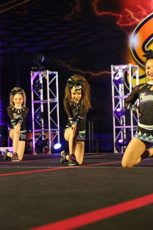 Young cheerleaders in matching black uniforms with hair bows kneel on a competition stage under bright lights.