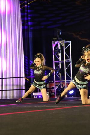Two young cheerleaders in sparkly uniforms strike energetic poses while kneeling on a stage with truss lighting.