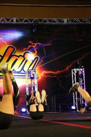 Cheerleaders lie on their backs with legs raised during a floor routine in front of a lightning-themed backdrop.