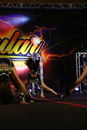 A cheerleader leaps with hair flying while teammates bend forward during a dynamic stage routine.