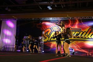 A cheer team performs a stunt sequence on a large competition floor with a glowing Spectacular banner behind them.