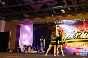 Cheerleaders lift a flyer into an extended stunt while teammates watch on a stage at The Spectacular cheer event.