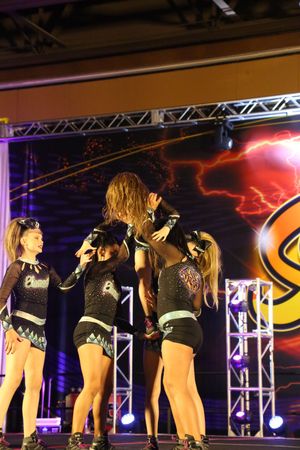 Cheer athletes in black and teal uniforms gather around a teammate during a stunt sequence on a lit competition stage.
