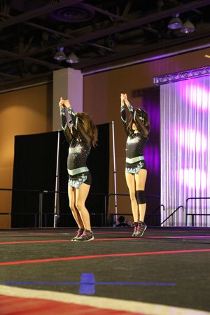 Two cheerleaders in matching black uniforms raise their arms in a synchronized pose on a competition floor with purple stage lighting.