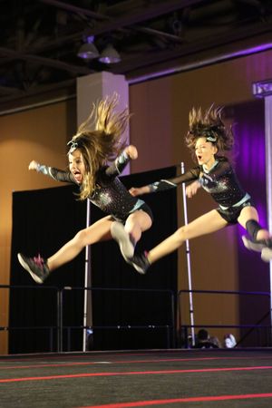 Two cheerleaders leap mid-air in matching black sparkly uniforms with hair flying during a dynamic competition routine.