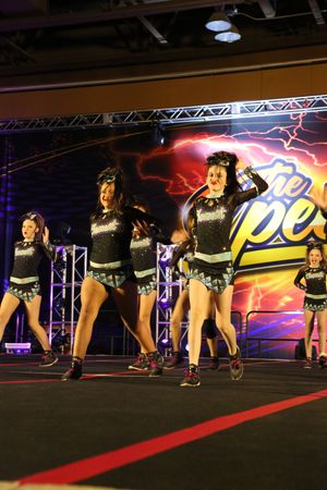 A cheer team in black uniforms performs a synchronized dance routine in front of a colorful lightning-themed Spectacular backdrop.