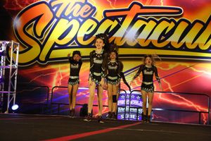 Cheerleaders in black sparkly uniforms land a jump together on a competition stage beneath a fiery Spectacular banner.