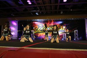 A cheer team in black uniforms kneels in formation on the competition floor in front of a lightning-themed Spectacular backdrop.