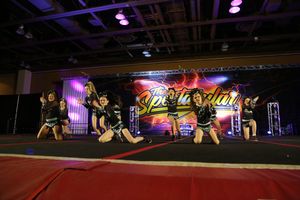 A cheerleading team poses in a dynamic formation on the mat at a competition with a colorful Spectacular banner behind them.