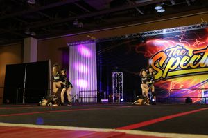 Cheer team performs on a competition stage under purple lights with The Spectacular banner glowing in the background.