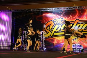 Cheerleaders in black uniforms perform a lifting stunt on stage in front of a colorful lightning-themed Spectacular backdrop.