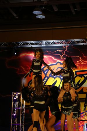 Youth cheerleaders in black uniforms perform a stunt with a flyer lifted high on stage in front of a lightning-themed backdrop.