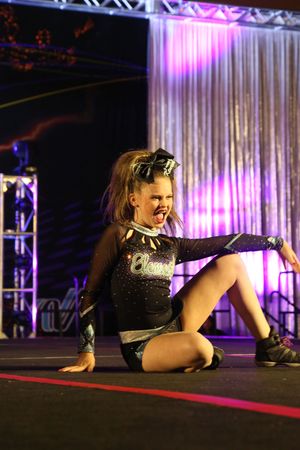 A young cheerleader with a large bow sits on the stage floor mid-performance, shouting with a fierce expression in a sparkly uniform.