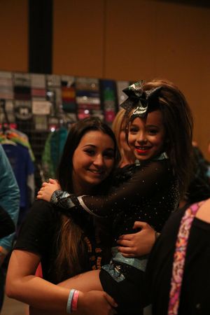 A smiling young cheerleader in a black uniform with a large bow is hugged by a teen girl at a cheer competition venue.