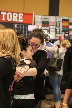 A woman with glasses hugs a young cheerleader in a sparkly black uniform at a busy cheer competition venue.