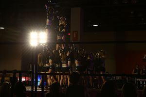 Silhouetted cheer team lifts a flyer high in a pyramid formation under bright stage lights at a competition.