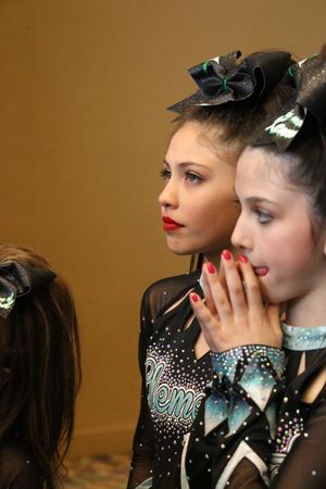 Two young cheerleaders in black Element uniforms with large bows watch anxiously, one covering her mouth with red nails.