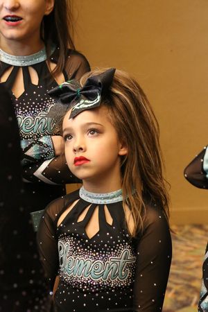 Young cheerleader with red lipstick and a large black bow gazes upward in an Element cheer uniform with crystal details.