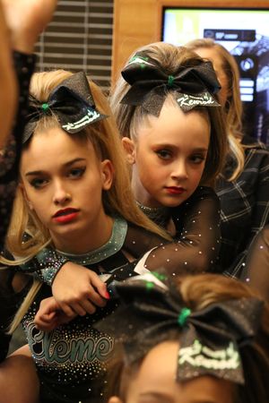 Two young cheerleaders in black Element uniforms and large glittery bows rest close together with serious expressions.