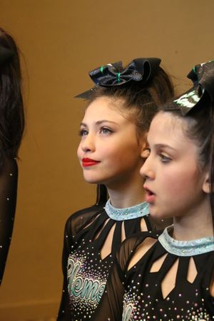 Two young cheerleaders with red lipstick and black bows in sparkly Element uniforms look off to the side at an event.