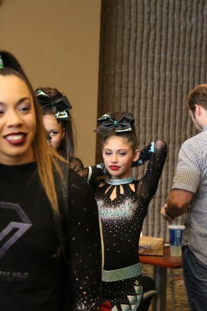 Smiling young cheerleader in a sparkly black and teal uniform adjusts her hair near teammates at a cheer competition.