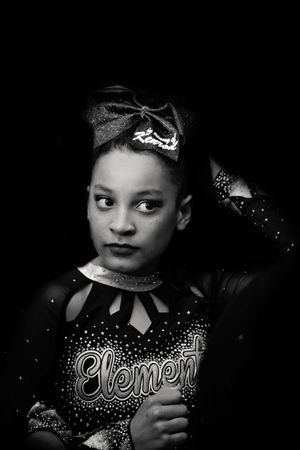Black and white portrait of a young cheerleader in an Element uniform with a sparkly bow glancing sideways.