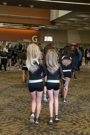 Two cheerleaders with long blonde hair in sparkly black uniforms walk through a crowded convention hall near the Hall G sign.
