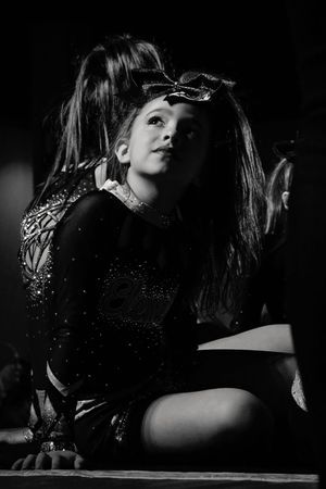 Black and white photo of a young cheerleader in a sparkly uniform and hair bow sitting on the floor and looking upward in dramatic shadows.