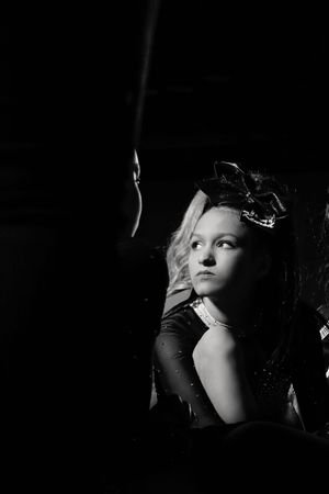 Black and white portrait of a young girl with wavy hair and a large bow resting her chin on her hand in dramatic low-key lighting.
