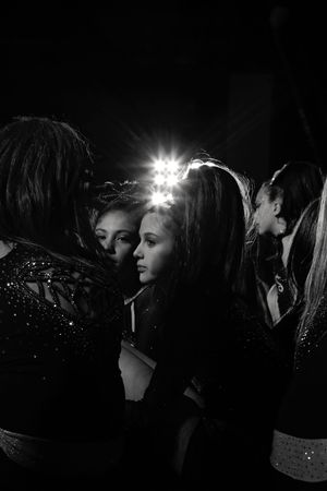 Black and white backstage photo of cheerleaders huddled together in sparkly uniforms with a bright stage light glowing behind them.