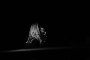 Moody black and white profile of a girl with long light hair emerging from deep darkness in a dramatic low-key portrait.