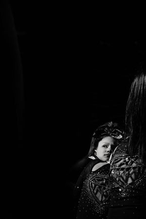 Black and white image of a young cheerleader with a hair bow glancing over her shoulder at a teammate in a sparkly uniform amid deep shadow.