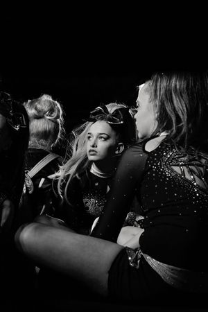 Black and white photo of a young cheerleader with a large bow gazing sideways, surrounded by teammates in sparkly competition uniforms.