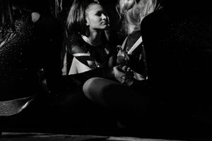 Dimly lit black and white photo of cheerleaders in sparkly uniforms sitting close together and talking quietly backstage.