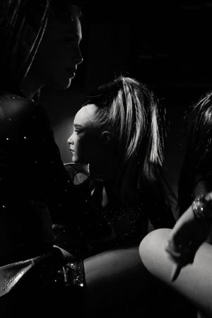 Black and white side view of a cheerleader with a high ponytail sitting between teammates in dramatic low light before competition.