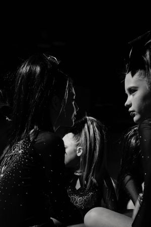 Black and white photo of young cheerleaders in sparkly uniforms gathered closely in dim light, two facing each other in profile.