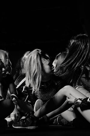 Black and white photo of a group of women sitting close together on a dark stage, two of them leaning in for an intimate kiss.