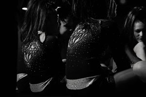 Black and white photo of a cheerleader in a sparkling rhinestone uniform reflected in a mirror, with another girl seated nearby.