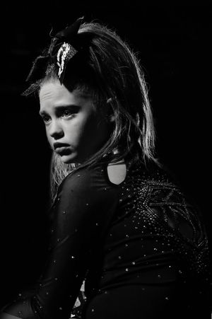 Black and white portrait of a young cheerleader in a sparkly long-sleeve uniform and large hair bow, gazing over her shoulder with a serious