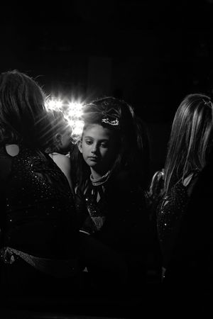 Black and white photo of a young girl wearing a tiara-style hair accessory gazing calmly while backstage lights glow behind her teammates.