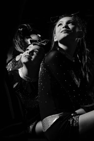 Black and white photo of two cheerleaders in sparkly uniforms and hair bows looking up hopefully, lit by dramatic light in a dark arena.