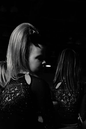 Black and white profile of a young cheerleader with a high ponytail and bow, wearing a sparkly rhinestone uniform in low light.