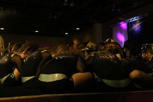 Cheerleading squad in black sequined uniforms gathers in a tight huddle on stage with purple lighting in the background.