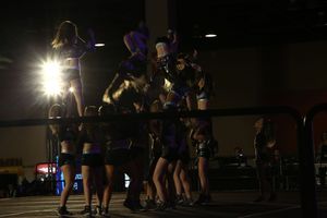 Cheerleaders in a dark arena lift a stunt group into the air while a bright spotlight glares behind them.