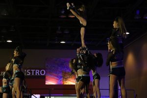 Cheerleader performs a high-flying stunt, balanced on one leg above her teammates, in a dimly lit competition arena.