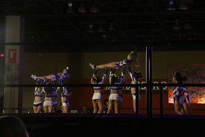 Cheer team in blue and white uniforms lifts a flyer horizontally in a stunt on a competition stage behind a railing.