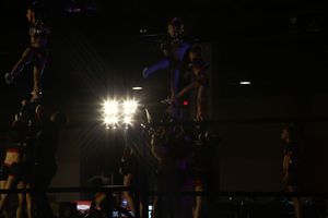 Silhouetted cheerleaders form a tall pyramid stunt in a dark arena with bright stage lights glowing behind them.