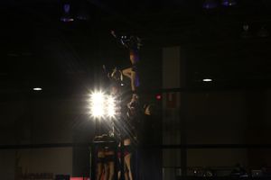 Cheerleader balanced high atop a stunt group is silhouetted against bright spotlights in a dark competition venue.